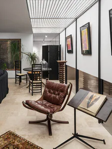 Modern gallery-style living area featuring a brown leather sling chair, black dining chairs, potted plants, framed abstract art, and a book titled Seeds & Fruits on a side table.