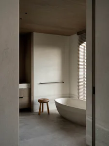 View into a modern minimalist bathroom with a concrete floor and walls, a white freestanding oval bathtub, a small three-legged oak stool, wooden cabinetry, and a window with wooden blinds.