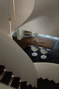 View from a curved white staircase looking down into a modern dining room with a long dark wood table, four white tulip chairs, and black marble flooring.