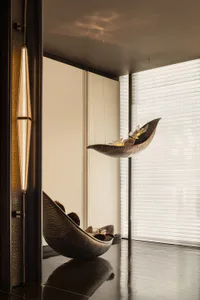 Modern luxury interior featuring a sculptural bronze leaf-shaped hanging lounge chair suspended from the ceiling and a matching floor version, with a tall pendant light on the left and large windows with sheer blinds in the background.