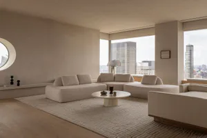 Modern minimalist living room with cream boucle modular sectional sofa, round travertine coffee table, large windows overlooking a city skyline, light wood flooring, and a circular wall mirror.
