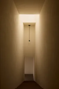 Looking up a narrow concrete staircase with a single black pendant light hanging from a high ceiling and warm indirect lighting from above.