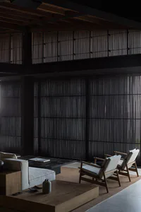 Modern minimalist living room featuring two wooden armchairs with light cushions, a low wooden coffee table, built-in platform seating, and dramatic floor-to-ceiling dark vertical wooden slat walls under a dark wood beamed ceiling.