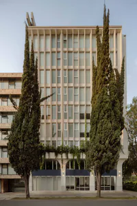 Front view of a multi-story brutalist concrete building with vertical window grids, arched ground floor entrances, climbing ivy, and two tall cypress trees on either side.