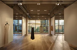 Spacious minimalist fashion boutique interior featuring exposed concrete pillars, wooden ceiling, light oak flooring, and clothing pieces hanging from ceiling rails with large city-view windows in the background.