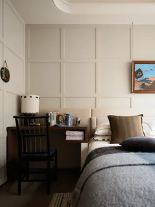 Modern neutral bedroom featuring a walnut desk, black spindle chair, upholstered bed with striped and textured pillows, table lamp, and framed landscape painting on a paneled wall.