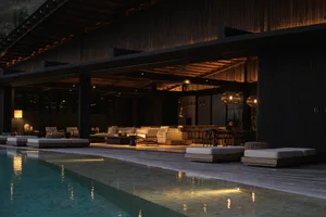 Night view of a modern luxury villa featuring an infinity pool, wooden deck, outdoor sofas, teak daybeds, woven rattan pendant lights, and a covered bar area with warm ambient lighting.