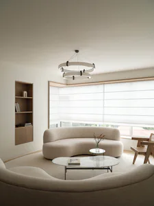 Minimalist living room featuring two curved boucle sofas, a round marble coffee table, a modern circular chandelier, built-in wooden shelving, and large windows with sheer roller blinds.