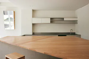 Modern minimalist kitchen featuring a large solid oak island with herringbone pattern, concrete base, white handleless cabinets, stainless steel floating shelves, and a stainless steel countertop with sink.