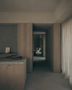 Modern minimalist interior hallway featuring floor-to-ceiling oak wood cabinetry, a concrete kitchen island with a wooden cutting board and fruit bowl in the foreground, textured concrete walls, and a view through to a distant living area with a light-colored armchair.