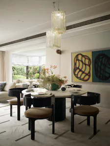 Modern dining room with a round white marble table, four black dining chairs with tan upholstery, a large sculptural glass chandelier, a vibrant floral centerpiece, and a large colorful abstract painting on the wall.