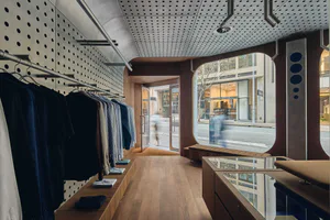 Interior view of a contemporary clothing boutique with walnut wood display fixtures, perforated metal ceiling, clothing racks, and large arched street-facing windows.
