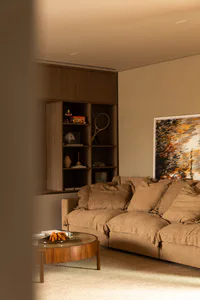 Modern living room interior with a large beige linen sectional sofa, round wooden coffee table, built-in wooden shelving unit displaying tennis racket, toys and decor, and an abstract painting on the wall.