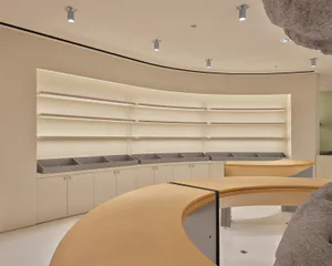 Modern minimalist retail interior featuring a large curved oak counter, built-in beige cabinetry with grey bins, and floating illuminated shelves under a curved ceiling with spotlights.