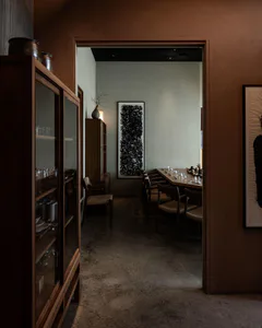 View through a doorway from a wooden glass cabinet into a minimalist Japanese-style dining room featuring terracotta walls, a long wooden communal table, woven-seat chairs, and large abstract black ink calligraphy artwork.