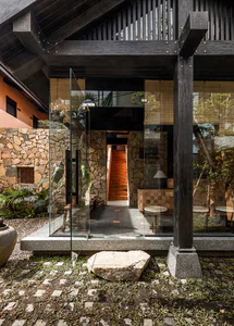 Modern tropical house entrance featuring large glass pivot doors, stacked stone walls, dark timber beams, and a view through to an orange staircase in the background.