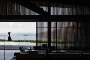 Modern open-plan living room interior with dark bamboo vertical screens, minimalist furniture, and a sweeping view of the ocean and distant rocky island through large openings.