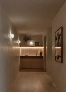 A narrow minimalist hallway with textured beige walls, a floating wooden console under a backlit shelf, spherical wall sconces, and a framed abstract artwork on the right.