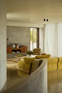 Modern living room interior with raw concrete walls, a curved concrete foreground element, mustard yellow boucle sofa, wooden media console with vinyl records, and large glass doors opening to greenery.