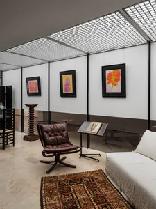 Modern gallery interior with white walls displaying three framed floral watercolor paintings, a brown leather lounge chair with wooden swivel base, a small black side table with a book, a patterned Persian-style rug, and a white sofa in the foreground under a grid skylight.