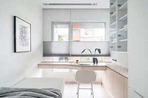 Modern minimalist home office featuring an L-shaped light wood desk, white ergonomic chair, built-in storage cabinets, large window with blinds, and a single bed in a bright, clean room.