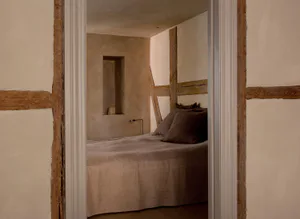 View through a doorway into a minimalist wabi-sabi style bedroom with exposed wooden beams, textured plaster walls, and a bed dressed in earthy brown linen bedding.