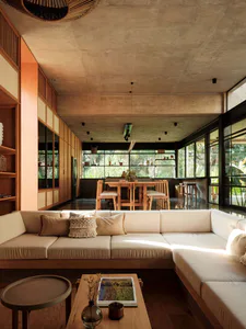 Modern tropical living room featuring a large off-white L-shaped sectional sofa on a wooden platform, concrete ceiling and walls, warm wood cabinetry, and a dining area with wooden table and chairs in the background, with lush greenery visible through large glass windows.