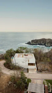 Aerial photograph of a modern white concrete villa with a curved infinity pool and rooftop terrace, dramatically situated on a rocky cliff overlooking the Pacific Ocean with lush tropical vegetation surrounding the property.