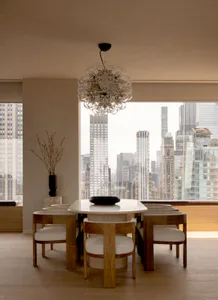 Modern dining room featuring a round white marble table, six oak and upholstered dining chairs, a large glass bubble chandelier, and floor-to-ceiling windows revealing a dense New York City skyline.