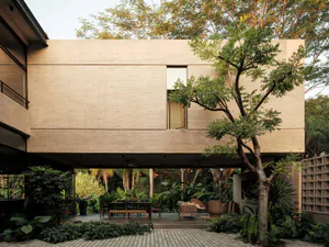 Modern concrete house with large open courtyard featuring tropical garden, outdoor dining area, and woven lounge furniture under a clear sky.