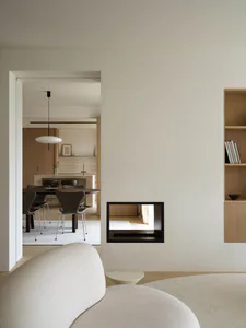 Modern minimalist living room with curved cream sofa, oak built-in shelving, and a black-framed pass-through fireplace offering a view into another room.