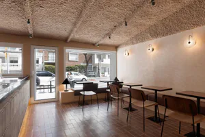 Interior view of a modern cafe with a heavily textured beige plaster ceiling, terracotta tile floor, built-in banquette seating, wooden chairs, and large windows overlooking a street scene.