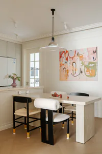 Modern dining room featuring a large travertine dining table, black dining chairs with white bouclé upholstery, a black pendant light, and a large colorful abstract painting on the wall.