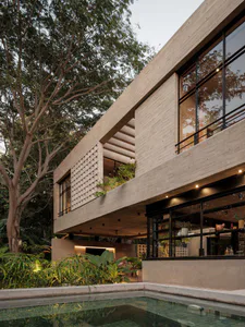 Modern two-story concrete house with black-framed windows, lattice screen, and cantilevered upper floor overlooking a reflecting pool surrounded by tropical plants at dusk.
