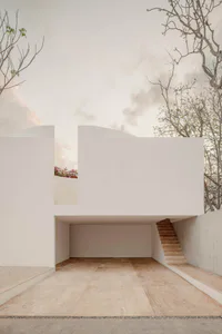 Modern minimalist concrete building with open courtyard, stone tile flooring, wooden staircase on the right, and trees against a cloudy sky.