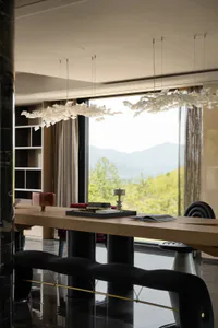 Modern dining room interior featuring a long oak table, black sculptural chairs, a black velvet bench with brass detail, and two large artistic chandeliers resembling flocks of white butterflies, with a panoramic view of green mountains through floor-to-ceiling windows.