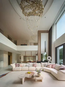 Modern double-height living room with a massive gold sculptural chandelier, curved white sectional sofa, marble coffee tables, and a second-floor mezzanine overlooking the space.