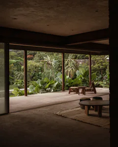 Modern tropical indoor-outdoor living space with polished concrete floors, wooden columns, low wooden furniture and a lush tropical garden visible through large openings.