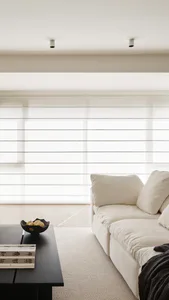 Modern minimalist living room with a white linen sectional sofa, black coffee table holding a dark bowl with fruit and books with Chinese characters, sheer horizontal blinds, and two recessed ceiling lights.