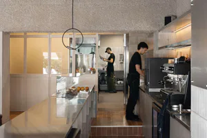 Modern industrial-style cafe kitchen featuring a speckled terrazzo ceiling, stainless steel counters, glass pastry display case, and two baristas working at espresso machines and prep stations.