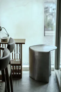 A modern curved concrete pedestal side table stands next to a wooden dining table with upholstered chairs and wine glasses, positioned by a large window with natural light.