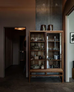 A tall oak display cabinet with glass doors filled with ceramic bowls, plates, and glassware, topped with two large brown pottery jars, standing in a dimly lit room with concrete walls and a hallway visible in the background.
