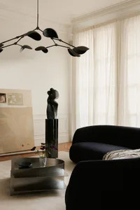 Modern living room interior featuring a large black velvet curved sofa, a multi-arm black kinetic chandelier, a dark abstract sculpture on a tall pedestal, a metallic tiered coffee table with purple flowers, and large abstract paintings leaning against a white wall with crown molding.