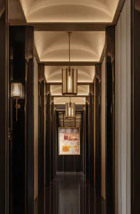 Long narrow luxury hotel hallway with dark wood paneling, arched ceilings with cove lighting, textured cylindrical pendant lanterns, and a colorful abstract painting at the end.