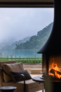 Modern lounge chair with beige cushions and striped pillow positioned beside a lit wood-burning stove, overlooking a misty green mountain and turquoise lake through large glass windows.