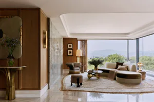 Modern luxury living room featuring a curved cream and brown sectional sofa, walnut wood paneling, marble walls, brass accents, and large floor-to-ceiling windows with mountain views.