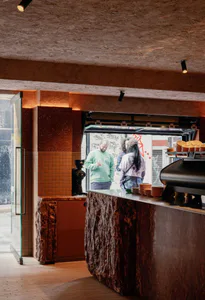 Interior view of a modern coffee shop with a dramatic service counter made from rough volcanic rock and polished terrazzo, featuring orange-brown tiled walls, a coffee grinder, stacked ceramic cups, and customers interacting at the window-style service hatch under warm spotlighting.