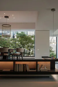 Modern minimalist dining room featuring a large window overlooking green trees, two mesh pendant lights, upholstered dining chairs, and a built-in illuminated travertine bookshelf filled with books in the foreground.