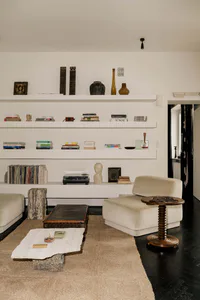 Modern minimalist living room with white built-in shelves displaying books and art objects, a large irregular travertine coffee table on a jute rug, a beige modular sofa, and a dark wood spool side table on dark flooring.