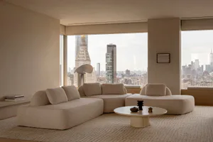 Modern minimalist living room with a large beige boucle sectional sofa, round travertine coffee table, and floor-to-ceiling windows overlooking the New York City skyline including the Empire State Building.